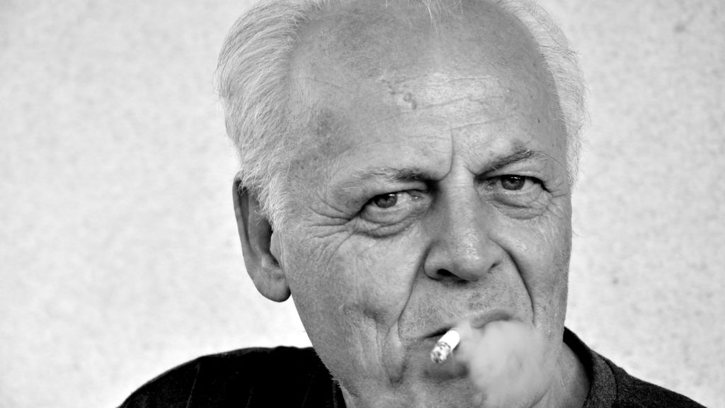 Starszy mężczyzna z papierosem w ustach ma zmarszczki palacza na twarzy.